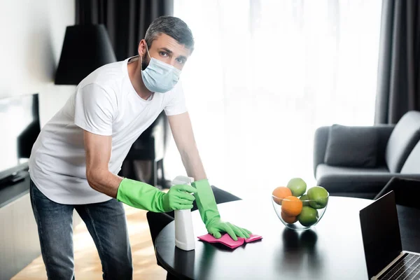 Mann mit medizinischer Maske und Gummihandschuhen hält Lappen und Waschmittel im Wohnzimmer — Stockfoto