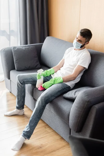 Selektiver Fokus des Mannes in medizinischer Maske, der Lappen und Waschmittel hält, während er auf der Couch sitzt — Stockfoto
