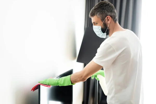 Side view of man in medical mask cleaning tv with rag and detergent in living room — Stock Photo