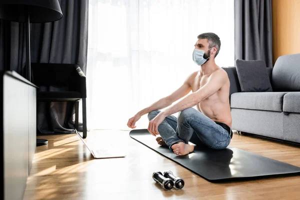 Vista lateral del hombre sin camisa en máscara médica sentado en la colchoneta de fitness cerca de pesas y portátil en casa - foto de stock