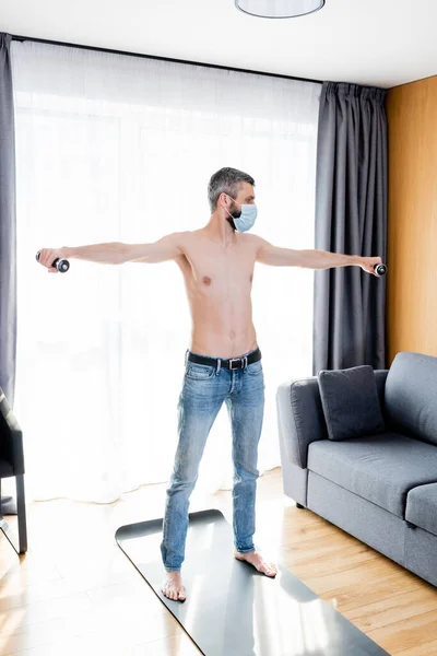 Shirtless man in medical mask working out with dumbbells in living room — Stock Photo