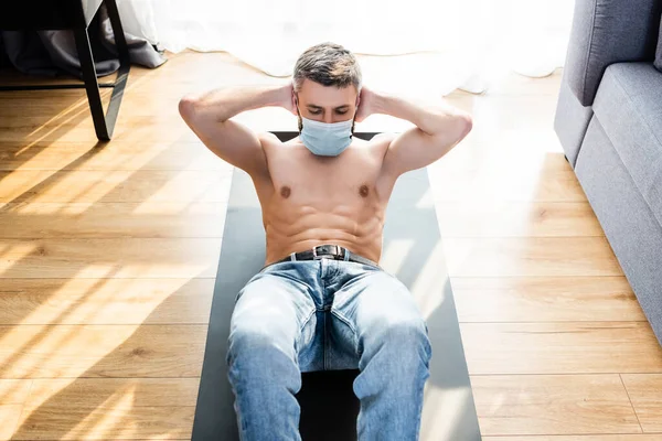 Vista de ángulo alto del hombre musculoso en la máscara médica haciendo abdominales mientras entrena en la estera de fitness en la sala de estar - foto de stock