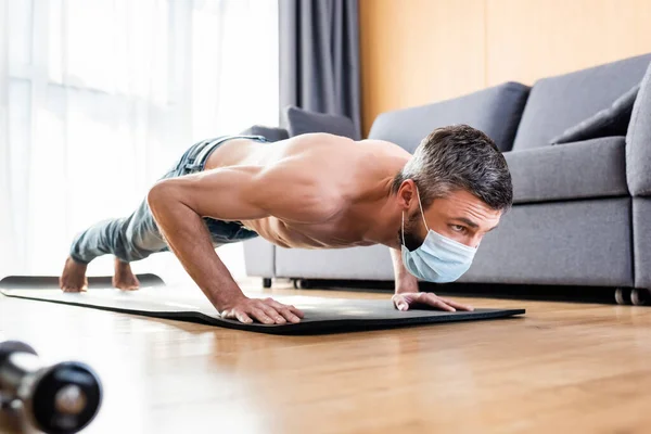 Foco seletivo do homem sem camisa na máscara médica fazendo press ups no tapete de fitness na sala de estar — Fotografia de Stock