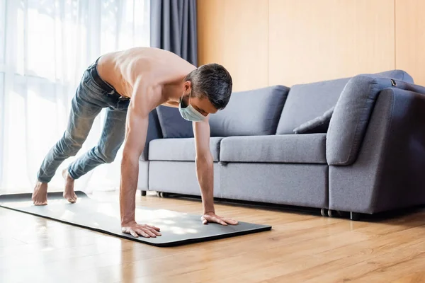 Hombre sin camisa con máscara médica haciendo ejercicio en la alfombra de fitness en casa - foto de stock