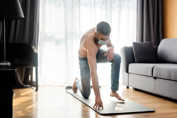 Homem muscular em máscara médica em pé no tapete de fitness em casa — Fotografia de Stock