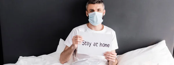 Colpo panoramico di uomo in maschera medica in possesso di carta con soggiorno a casa lettering sul letto — Foto stock