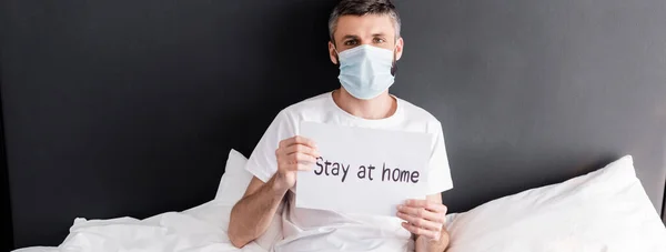 Tiro panorâmico de homem em cartão de retenção máscara médica com estadia em casa lettering na cama — Fotografia de Stock