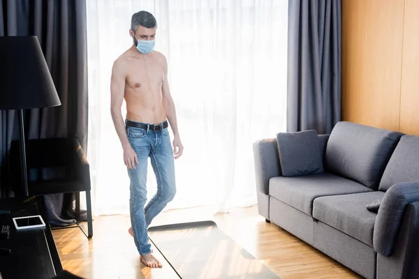Homem muscular em máscara médica em pé perto do tapete de fitness na sala de estar — Fotografia de Stock