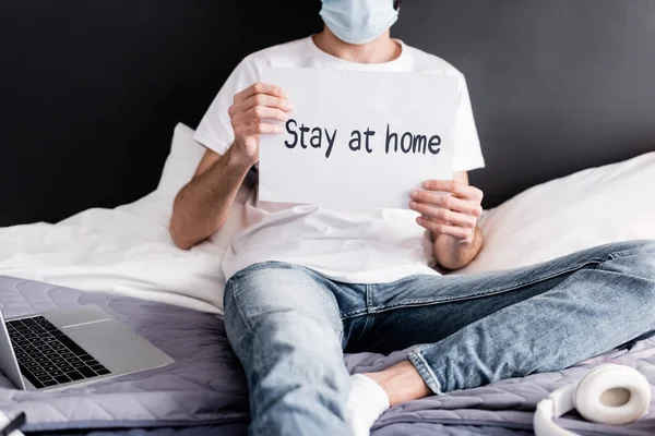 Vista cortada do homem em cartão de retenção máscara médica com estadia em casa lettering perto de laptop e fones de ouvido na cama — Fotografia de Stock