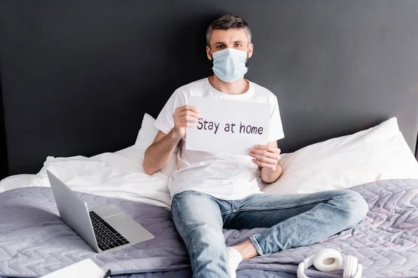 Homme en masque médical tenant la carte avec rester à la maison lettrage près de l'ordinateur portable et écouteurs sur le lit — Photo de stock