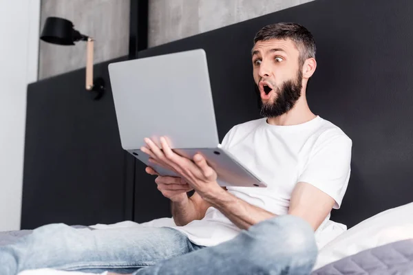 Selective focus of shocked man holding laptop on bed — Stock Photo