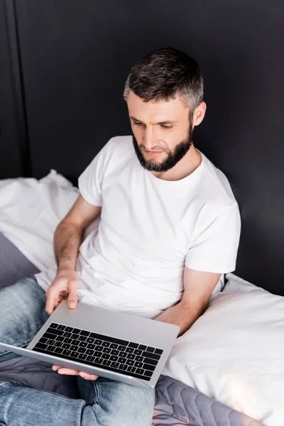 Guapo freelancer sosteniendo portátil mientras está sentado en la cama - foto de stock