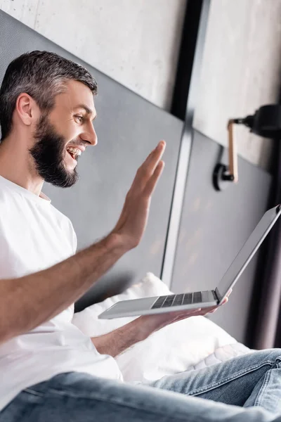 Vue latérale d'un homme souriant ayant un appel vidéo sur un ordinateur portable au lit — Photo de stock