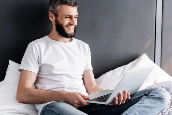 Guapo freelancer sonriendo mientras sostiene el ordenador portátil en la cama - foto de stock