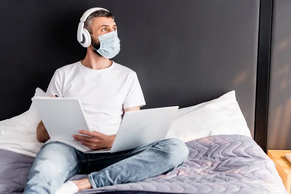 Freelancer en máscara médica y auriculares que trabajan con portátil y portátil en la cama - foto de stock
