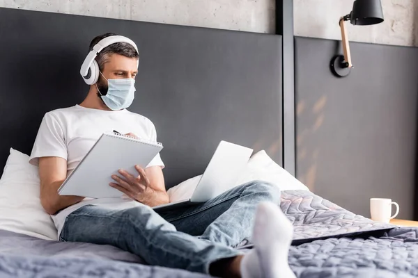 Enfoque selectivo del teletrabajador en auriculares y mascarilla médica blanqueando en el portátil y utilizando el ordenador portátil en el dormitorio - foto de stock