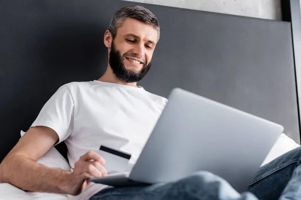 Selective focus of smiling freelancer holding credit card while using laptop on bed — Stock Photo