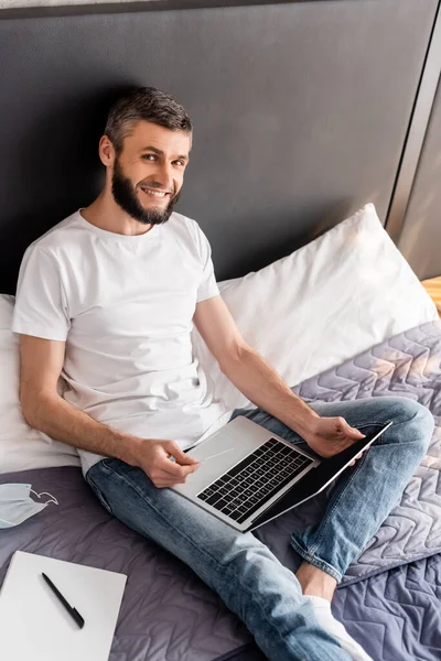 Hochwinkelaufnahme eines lächelnden Mannes mit Kreditkarte und Laptop in der Nähe medizinischer Maske und Notizbuch auf dem Bett — Stockfoto