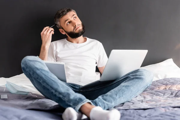Selective focus of pensive freelancer looking away while holding laptop and notebook on bed — Stock Photo