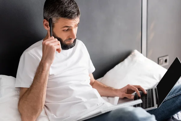 Selective focus of handsome teleworker working with laptop near notebook on bed — Stock Photo