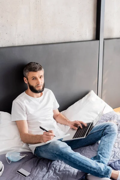 Vista ad alto angolo di freelance utilizzando laptop e scrittura su notebook vicino alla maschera medica e carta di credito sul letto — Foto stock