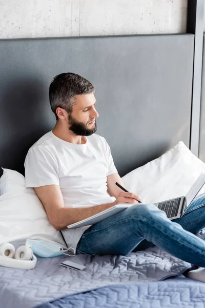 Teletrabajo guapo usando el ordenador portátil y escribir en el cuaderno cerca de la máscara médica y tarjeta de crédito en la cama — Stock Photo