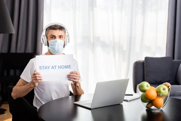 Teletrabajador con máscara médica y auriculares con tarjeta de retención con letras en casa cerca de la computadora portátil en la mesa - foto de stock