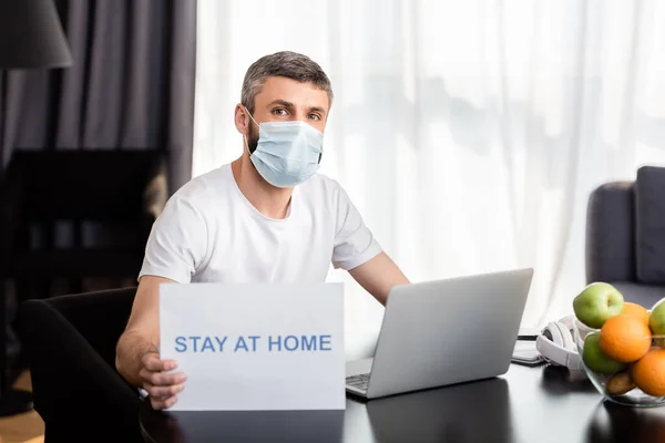 Freelancer en tarjeta de retención de máscara médica con letras en casa cerca de la computadora portátil y frutas en la mesa - foto de stock