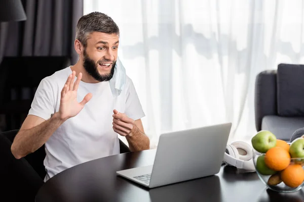 Freelance sorridente che fa videochiamata sul computer portatile e tiene la maschera medica a casa — Foto stock