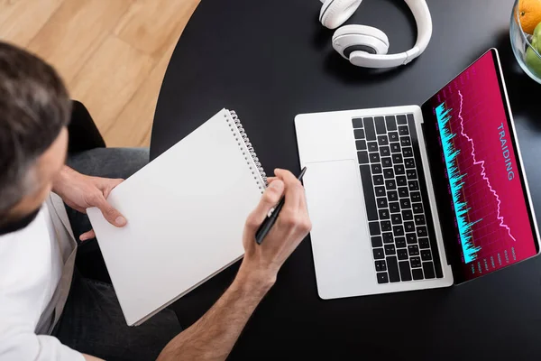 Overhead view of teleworker holding notebook and pen near laptop with graphs on screen — Stock Photo