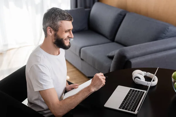 Vista lateral del freelancer sonriente sosteniendo portátil cerca de la computadora portátil y auriculares en la mesa en casa - foto de stock