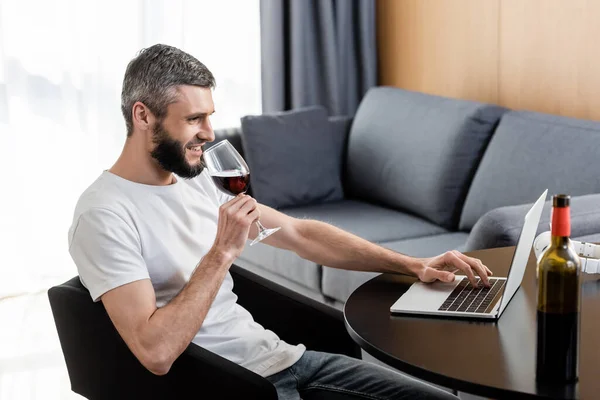 Vista lateral do sorrindo freelancer beber vinho e usando laptop na sala de estar — Fotografia de Stock