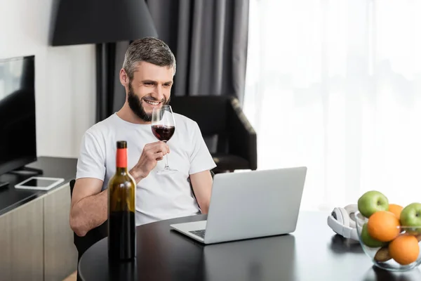Foco seletivo de sorrir freelancer segurando copo de vinho perto de laptop e frutas na mesa — Fotografia de Stock