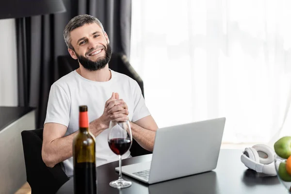 Enfoque selectivo del freelancer sonriente mirando la cámara cerca del vino y el portátil en la mesa en la sala de estar - foto de stock