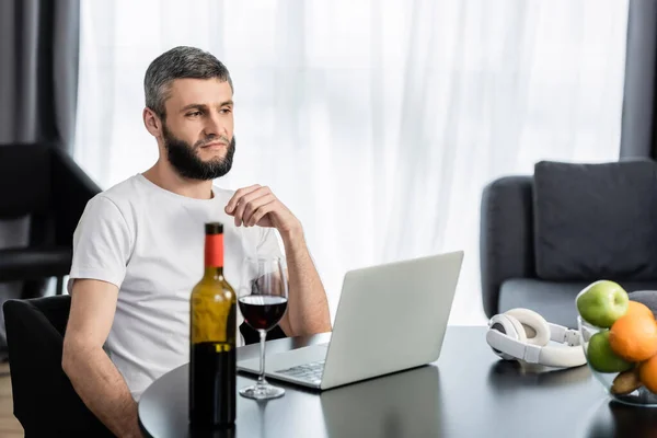 Selective focus of teleworker looking at laptop near wine and fruits on table — Stock Photo