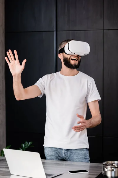 Hombre sonriente en auriculares vr jugando videojuego cerca de la computadora portátil y el teléfono inteligente en la mesa - foto de stock