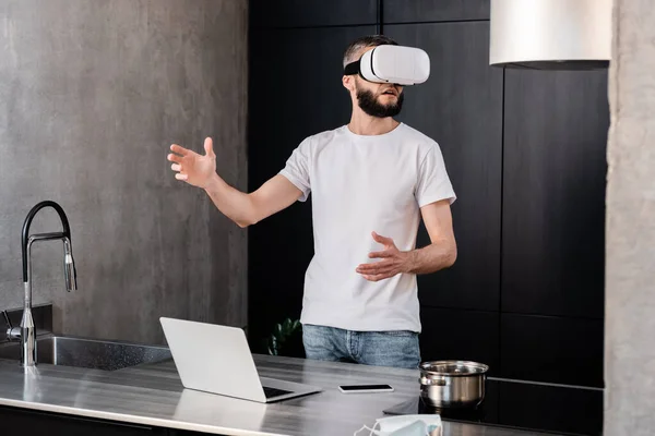 Focus selettivo dell'uomo utilizzando auricolare vr vicino gadget e maschera medica sul piano di lavoro della cucina — Foto stock