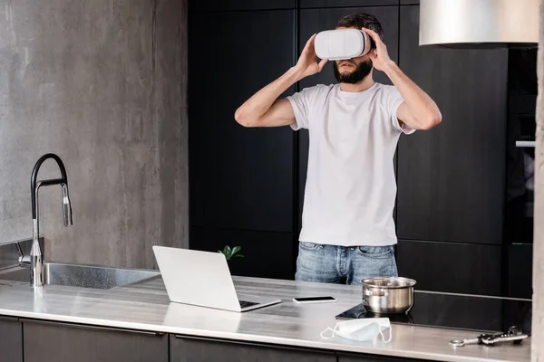 Uomo che utilizza auricolare realtà virtuale vicino a dispositivi digitali e maschera medica sul piano di lavoro della cucina — Foto stock