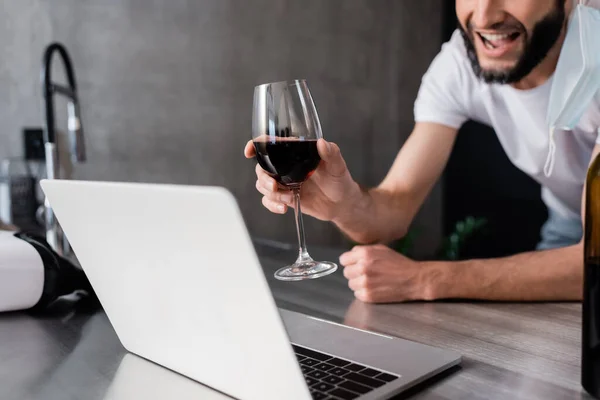 Vista recortada del hombre sonriente en máscara médica sosteniendo vaso de vino cerca de la computadora portátil en la encimera de la cocina - foto de stock