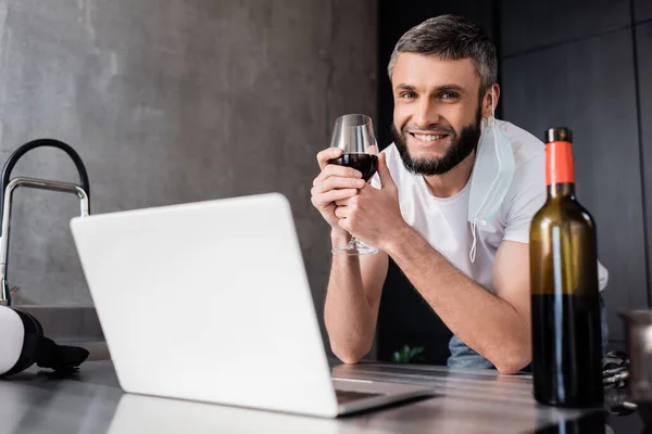 Foco seletivo de homem sorridente em máscara médica segurando vidro de vinho perto de laptop e fone de ouvido vr na bancada da cozinha — Fotografia de Stock