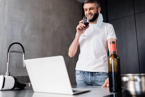 Enfoque selectivo del freelancer sonriente en máscara médica bebiendo vino cerca de la computadora portátil en la encimera en la cocina - foto de stock