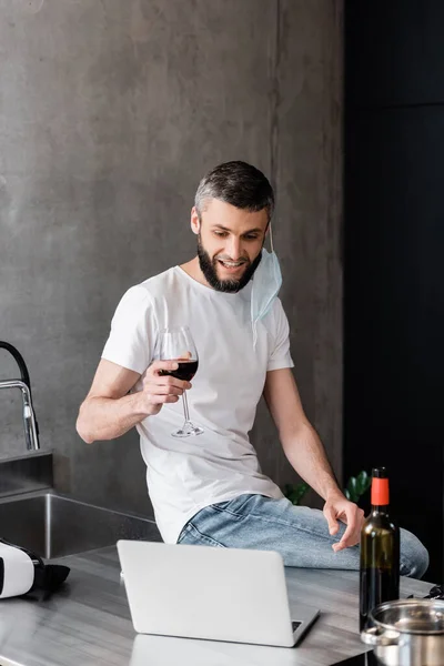 Hombre sonriente con máscara médica sosteniendo un vaso de vino cerca de la computadora portátil y auriculares vr en la encimera en la cocina - foto de stock