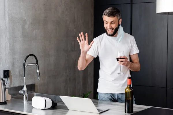 Homme souriant dans un masque médical tenant un verre de vin tout en ayant un appel vidéo sur un ordinateur portable dans la cuisine — Photo de stock