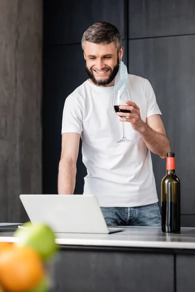 Foco seletivo de homem sorridente em máscara médica segurando copo de vinho e usando laptop na cozinha — Fotografia de Stock