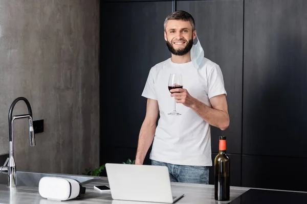 Homem em máscara médica segurando copo de vinho e sorrindo para a câmera perto do laptop na bancada na cozinha — Fotografia de Stock