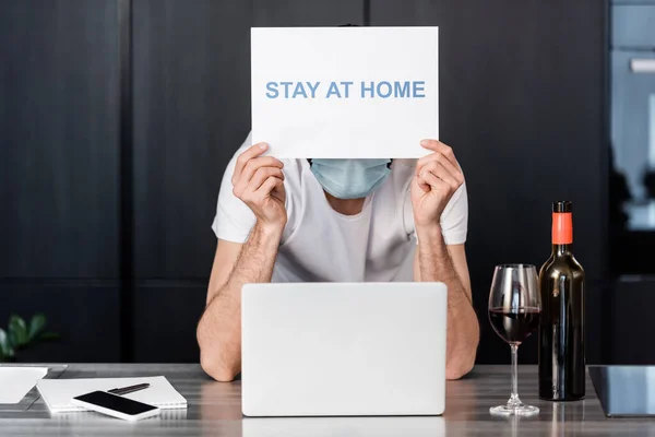 Homme en masque médical tenant la carte avec rester à la maison lettrage près de l'ordinateur portable, vin et ordinateur portable dans la cuisine — Photo de stock