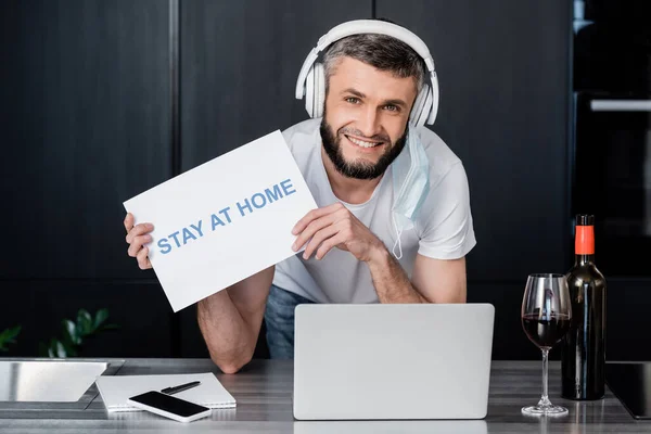 Freelancer sonriente en auriculares y tarjeta de retención de máscara médica con letras de estancia en casa cerca de gadgets y vino en la cocina - foto de stock