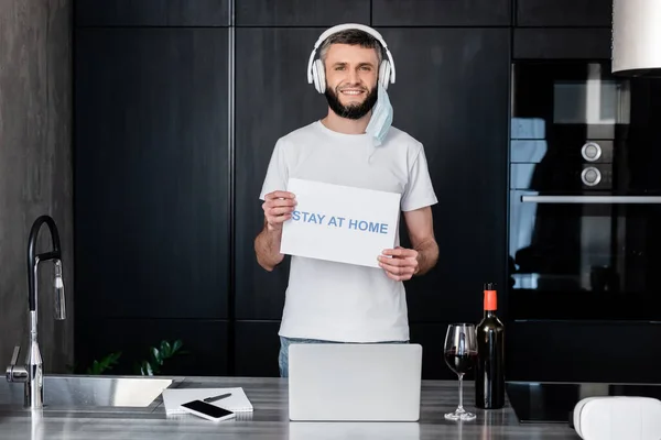 Uomo sorridente in cuffia e maschera medica con carta di soggiorno a casa lettering vicino al vino e dispositivi digitali sul piano di lavoro della cucina — Foto stock