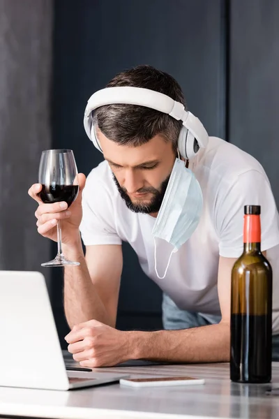 Foco seletivo de freelancer em máscara médica e fones de ouvido segurando copo de vinho perto de gadgets na bancada na cozinha — Fotografia de Stock