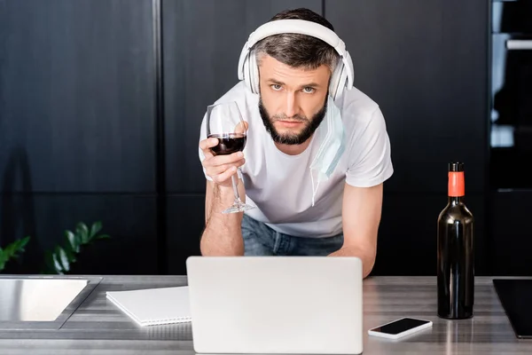 Handsome freelancer in headphones and medical mask looking at camera while holding glass of wine near gadgets on worktop in kitchen — Stock Photo